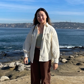 Mundra is wearing a cream jacket and standing in front of the ocean