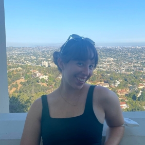Emma is wering a black shirt and is standing in front of a window with a view of buildings and hills below
