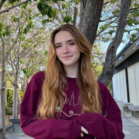 Frannie is wearing a Burgundy sweatshirt and is standing on a walkway with several trees in the background