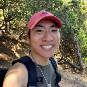 Nathan is wearing a black shirt, backpack and a red hat with trees in the background 