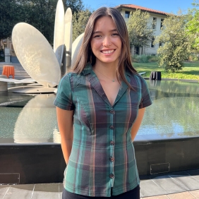 Sophia is wearing a green plaid shirt and is standing in front of a fountain at the Oxy Campus